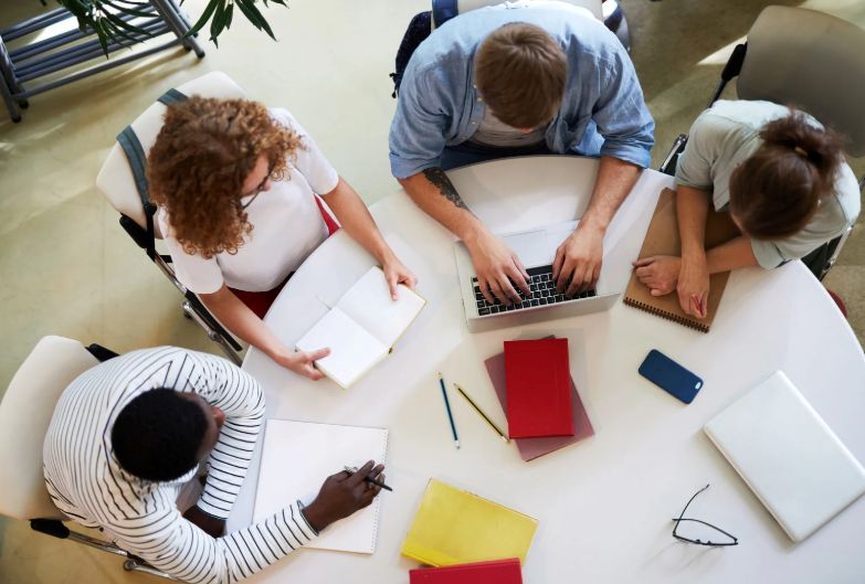 Vier Personen sitzen an einem runden Tisch und arbeiten mit Laptop, Notizbüchern und Stiften.